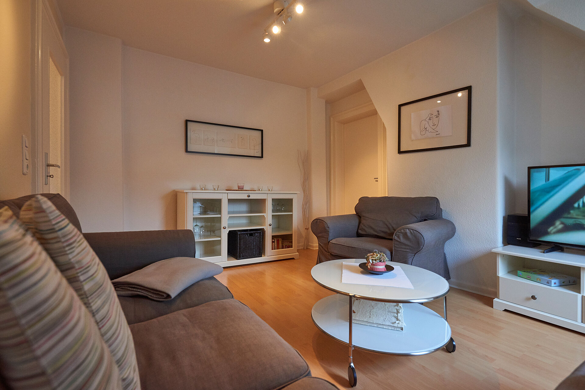 Wohnzimmer mit Couch, Sessel, Sideboard und TV, der weiße Couchtisch steht auf Rollen
