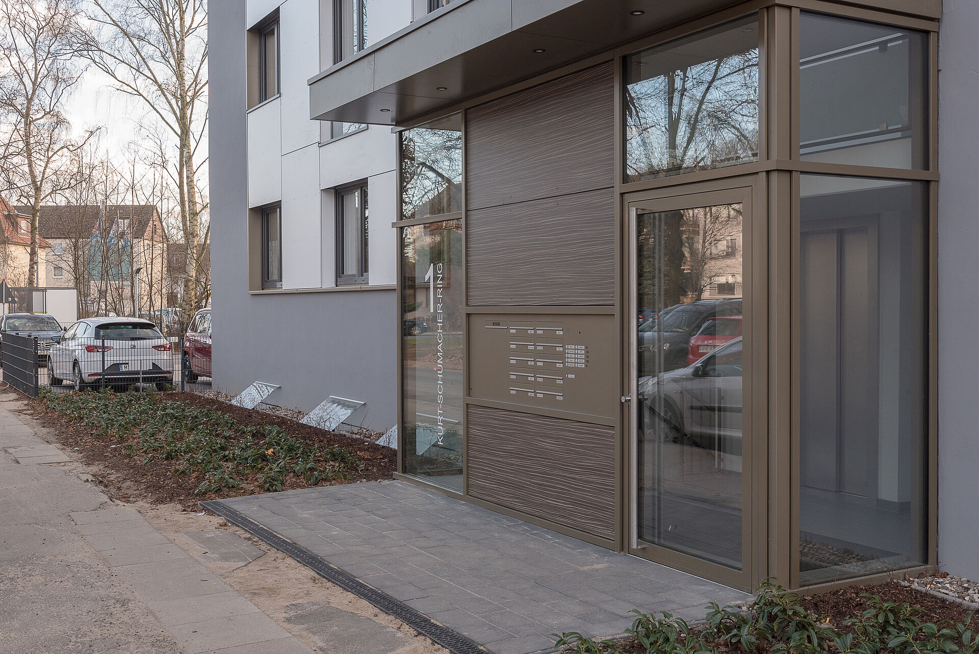 Gepflegter Hauseingang mit großen Glasfronten und kleinen grünen Beeten vor der Tür