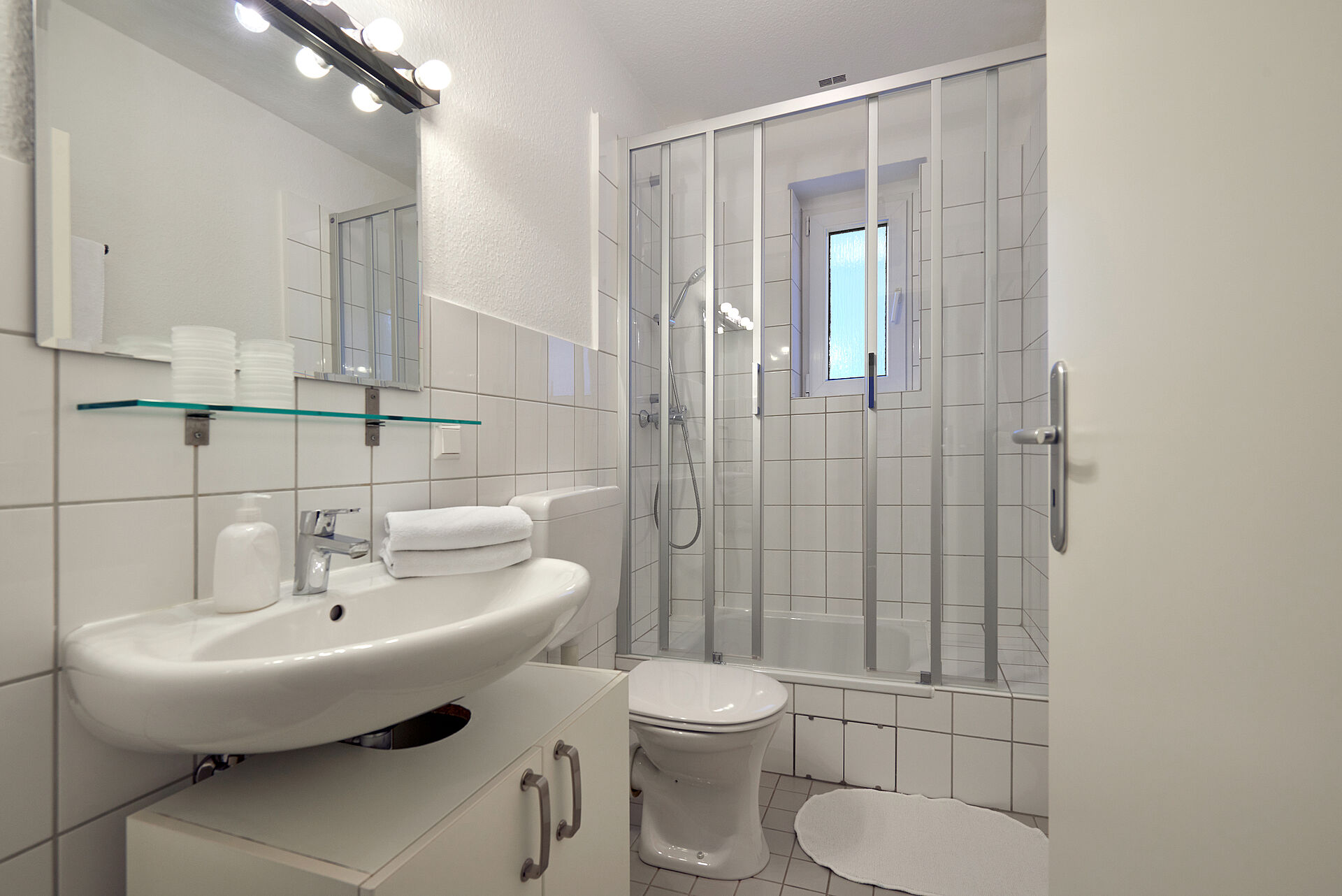 Badezimmer mit Dusche und einem beleuchteten Spiegel, auf dem Waschbecken liegen frische Handtücher