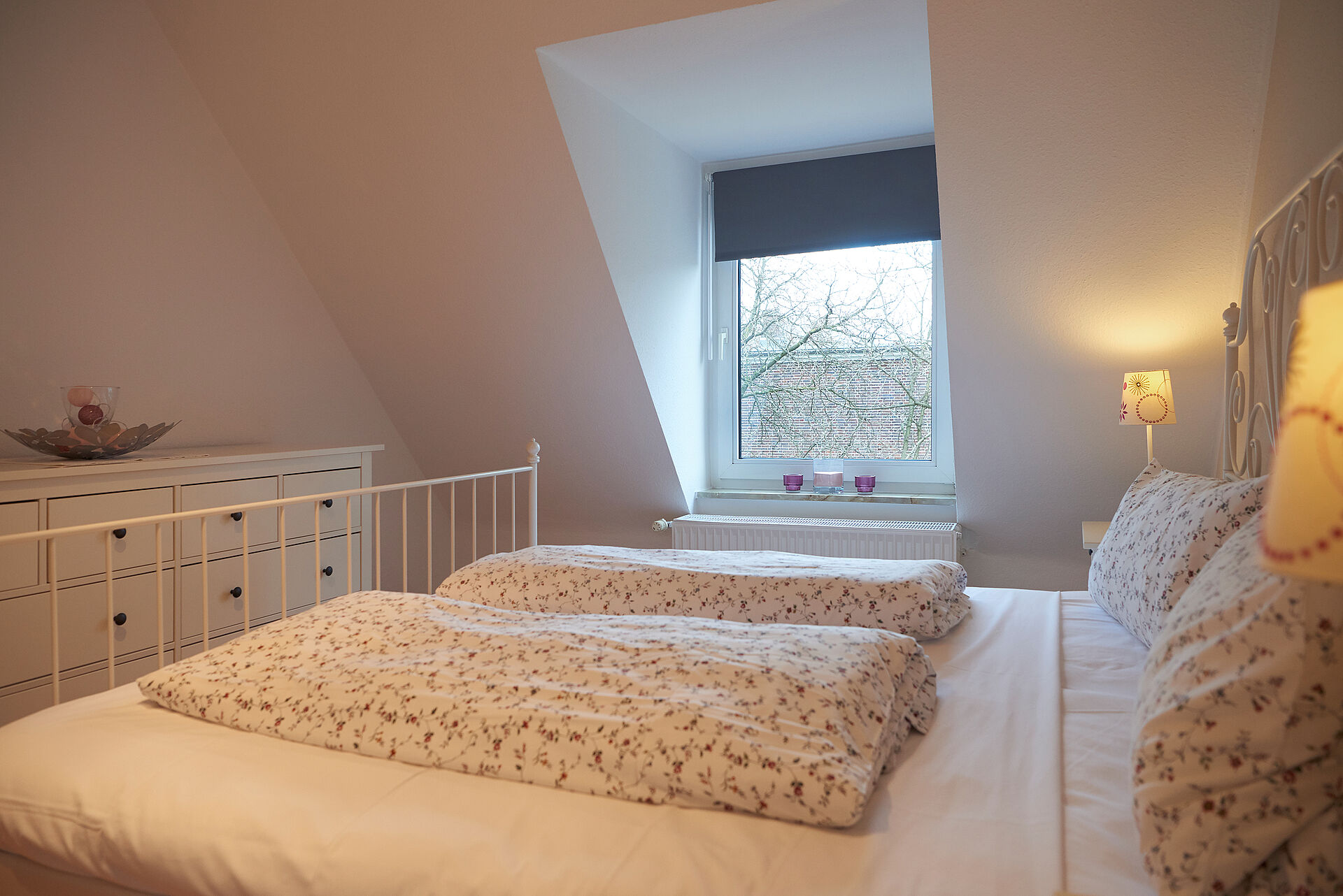 Blick über ein frisch bezogenes Bett auf eine Kommode und das Schlafzimmerfenster