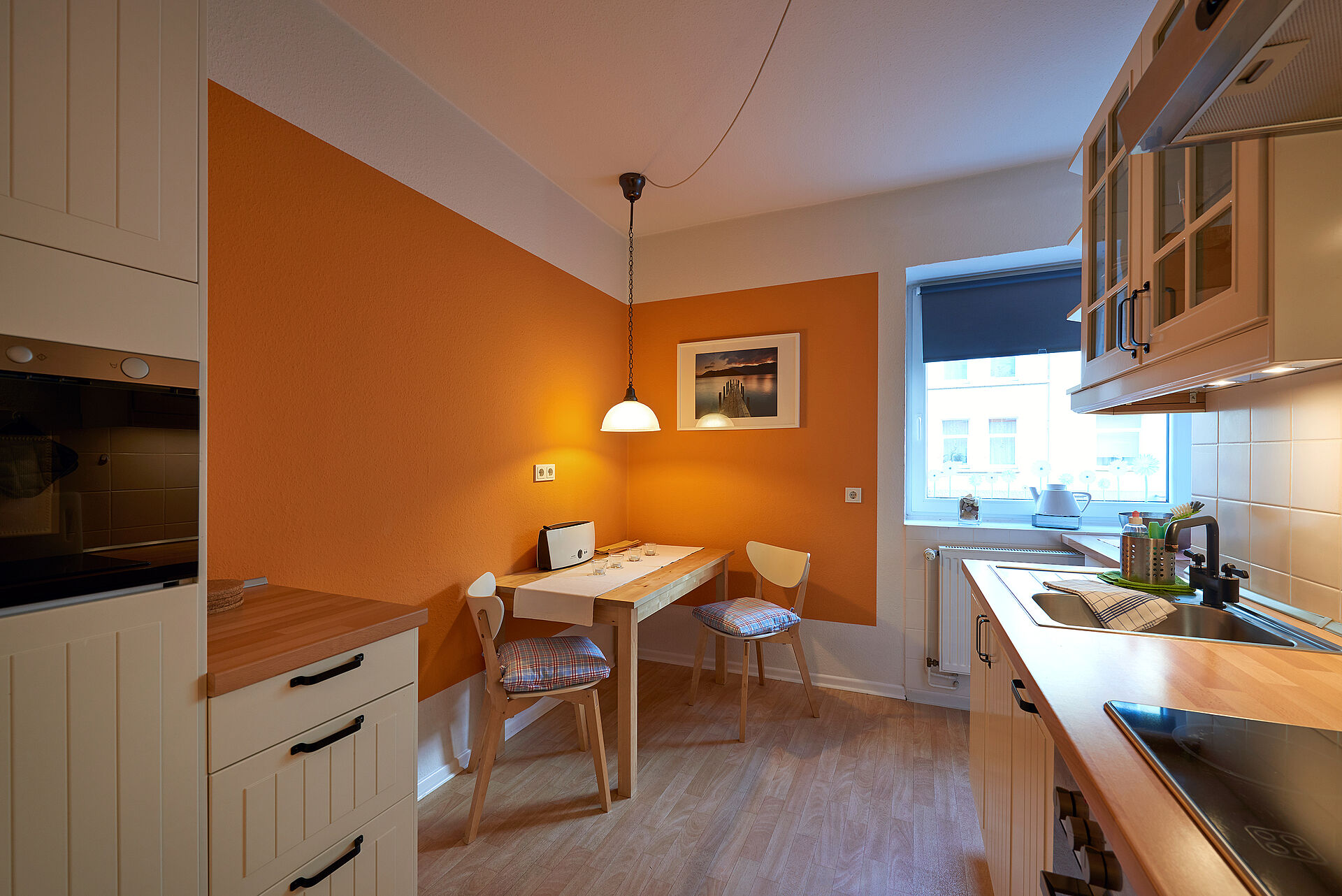 Eine helle Küche mit einer orange gestrichenen Wand, davor ein Küchentisch mit zwei Stühlen